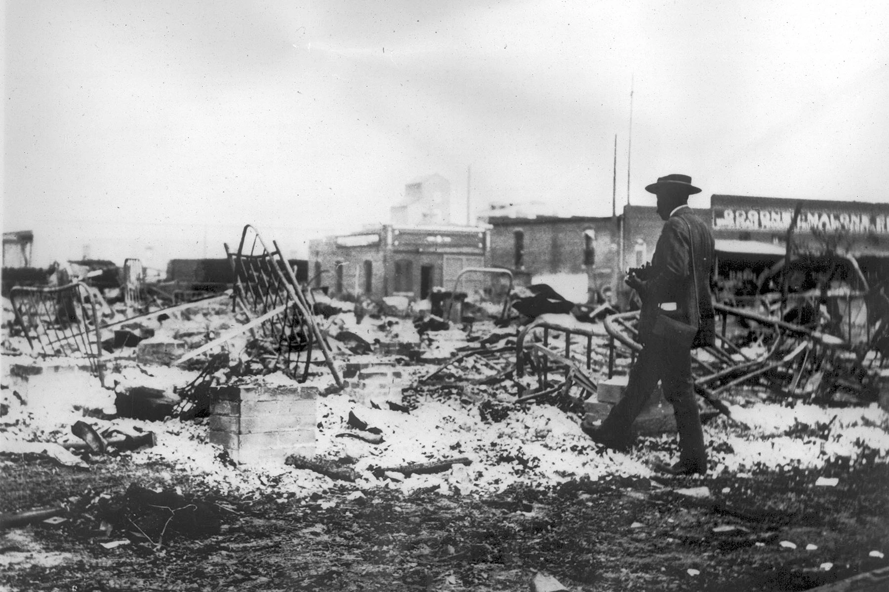 A black man with a camera looks at the skeletons of iron beds which rise above the ashes of a burned-out block after the Tulsa Massacre.