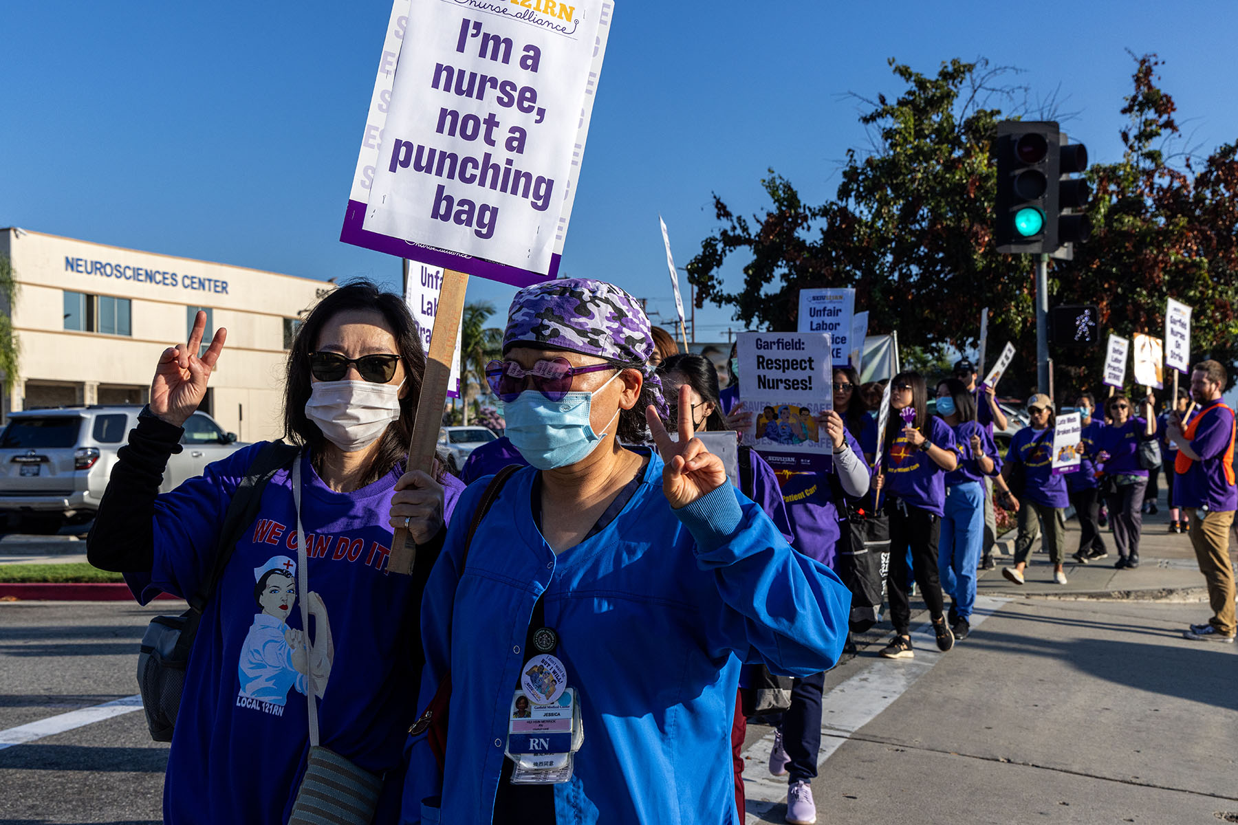 Photograph of two nurses standing in a line of protesters, both wearing masks and sunglasses. One nurse holds up a sign that says, “I’m a nurse, not a punching bag.”