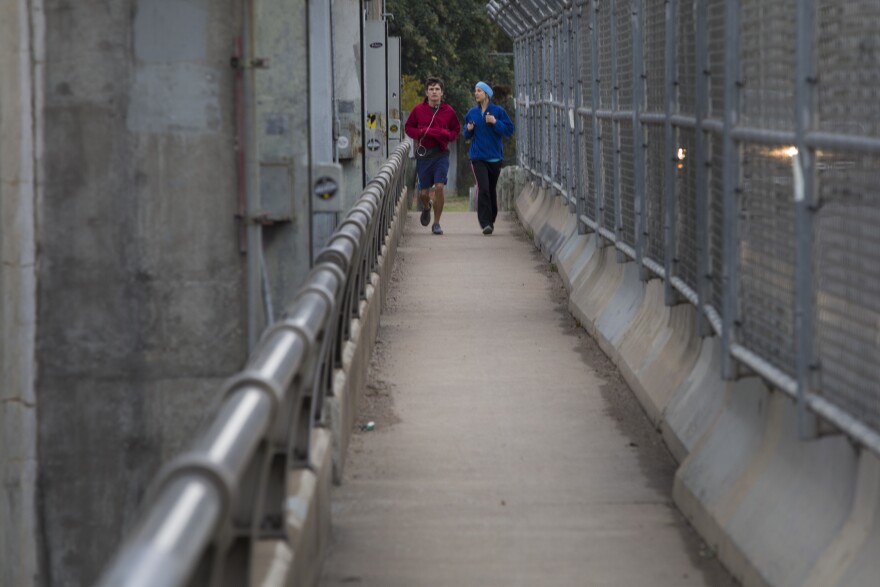 Two people are running along a narrow sidewalk over a bridge. Chainlink fence separates pedestrians from cars. A low guard rail was the only thing prevent people from falling in the lake.