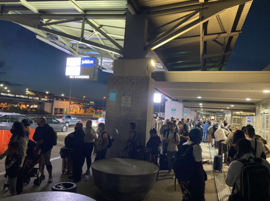 TSA security lines stretched onto A picture taken on a phone showing people with baggage standing in line outside the Barbra Jordan Terminal. The sky looks dark as if it's not even dawn yet. the sidewalk early Saturday morning at Austin-Bergstrom International Airport.