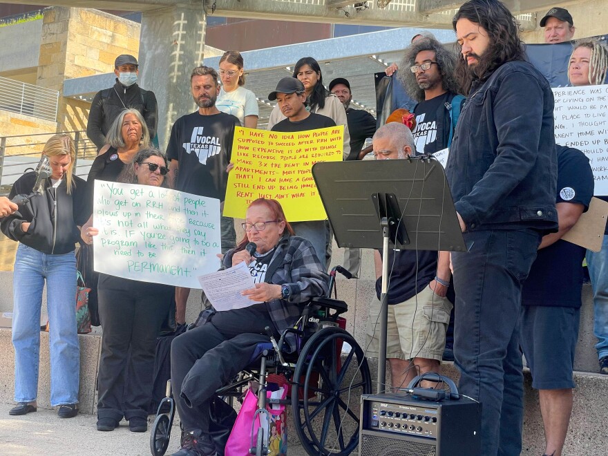 A woman with red hair and sitting in a wheel chair holding a white paper and microphone address the crowd. She is surrounded by many people holding signs and all wearing shirts that say Vocal Texas. 