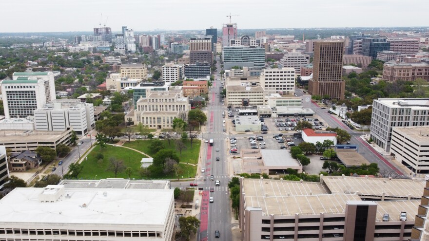 An aerial view of Guadalupe Street in March 2023. A view looking north from about 7th Street. A red stripe indicating a bus lane is visible on the road. Woolridge Square Park is seen as a small green field. Buildings line either side of the road. 